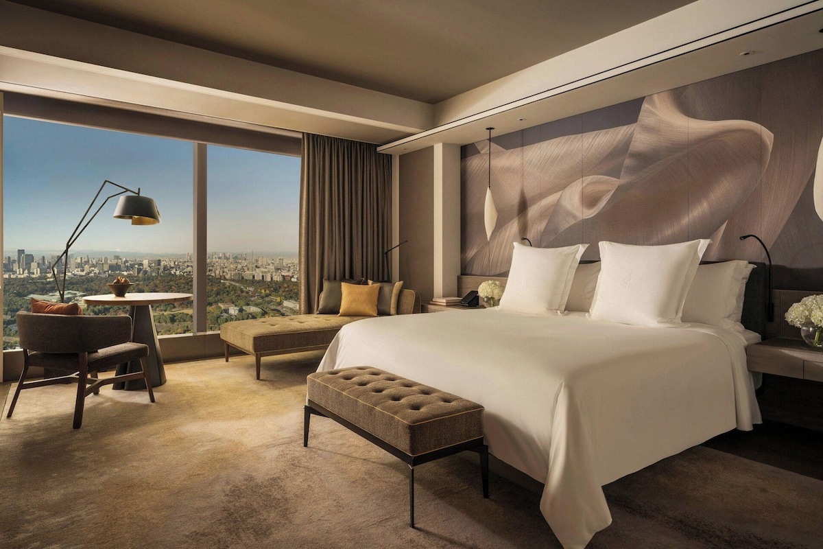 A modern hotel room with a large bed, a sitting area with a view, and a cityscape visible through the window.