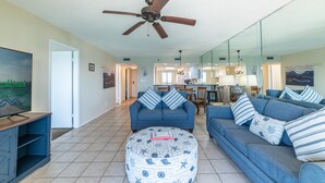A living room with a blue sofa, a round ottoman with nautical patterns, a ceiling fan, and a television on a wooden stand. - Rest Ashored Commodore 1303-Beachfront Master-Rest Assured, You'll Love It Here! (Panama City Beach)