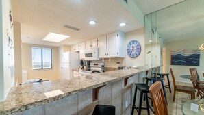 A modern kitchen with granite countertops, white cabinets, and a dining area with a glass table and chairs. - Rest Ashored Commodore 1303-Beachfront Master-Rest Assured, You'll Love It Here! (Panama City Beach)