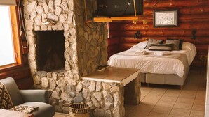 A cozy log cabin room with a stone fireplace, a bed with white bedding, a small table, and a sofa. - Ruma Andina by DOT Cabana (Villa La Angostura)