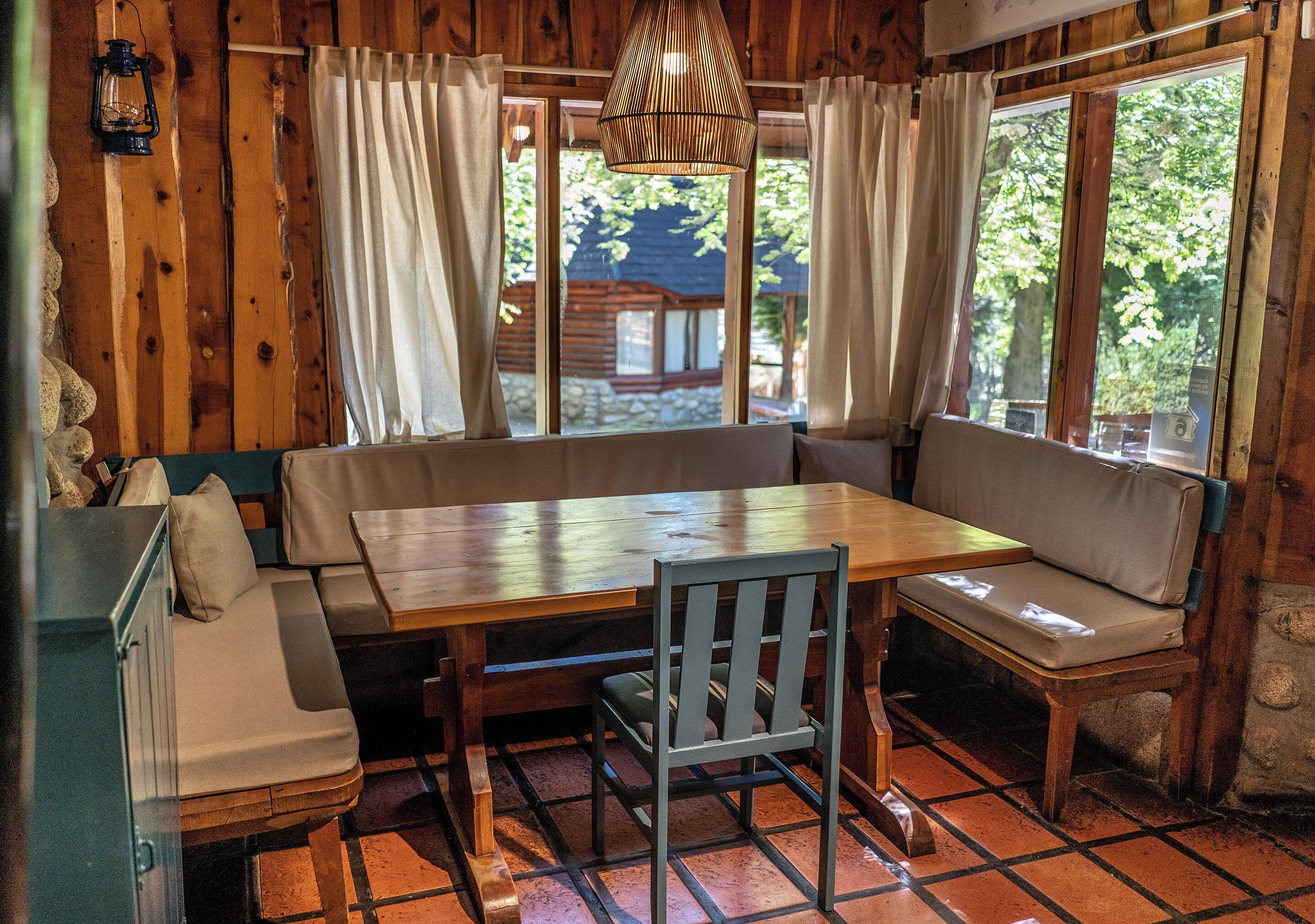 A wooden cabin interior with a dining table, bench seating, and a view of the outdoors.