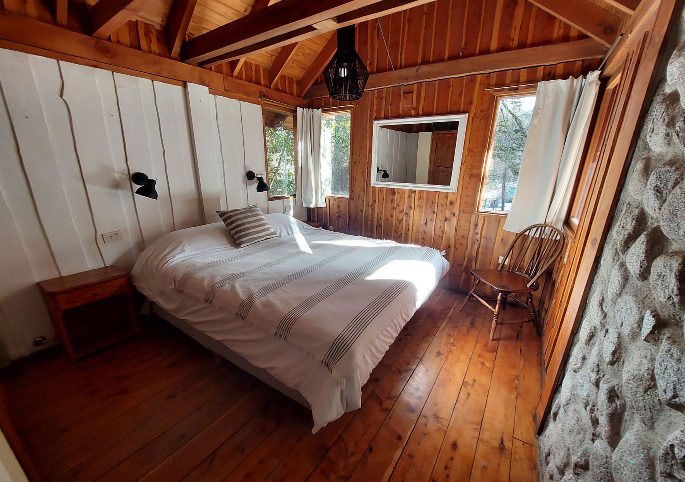 Une chambre dans une cabane en bois, avec un lit, une table de chevet, une chaise, un miroir et un mur en pierre.