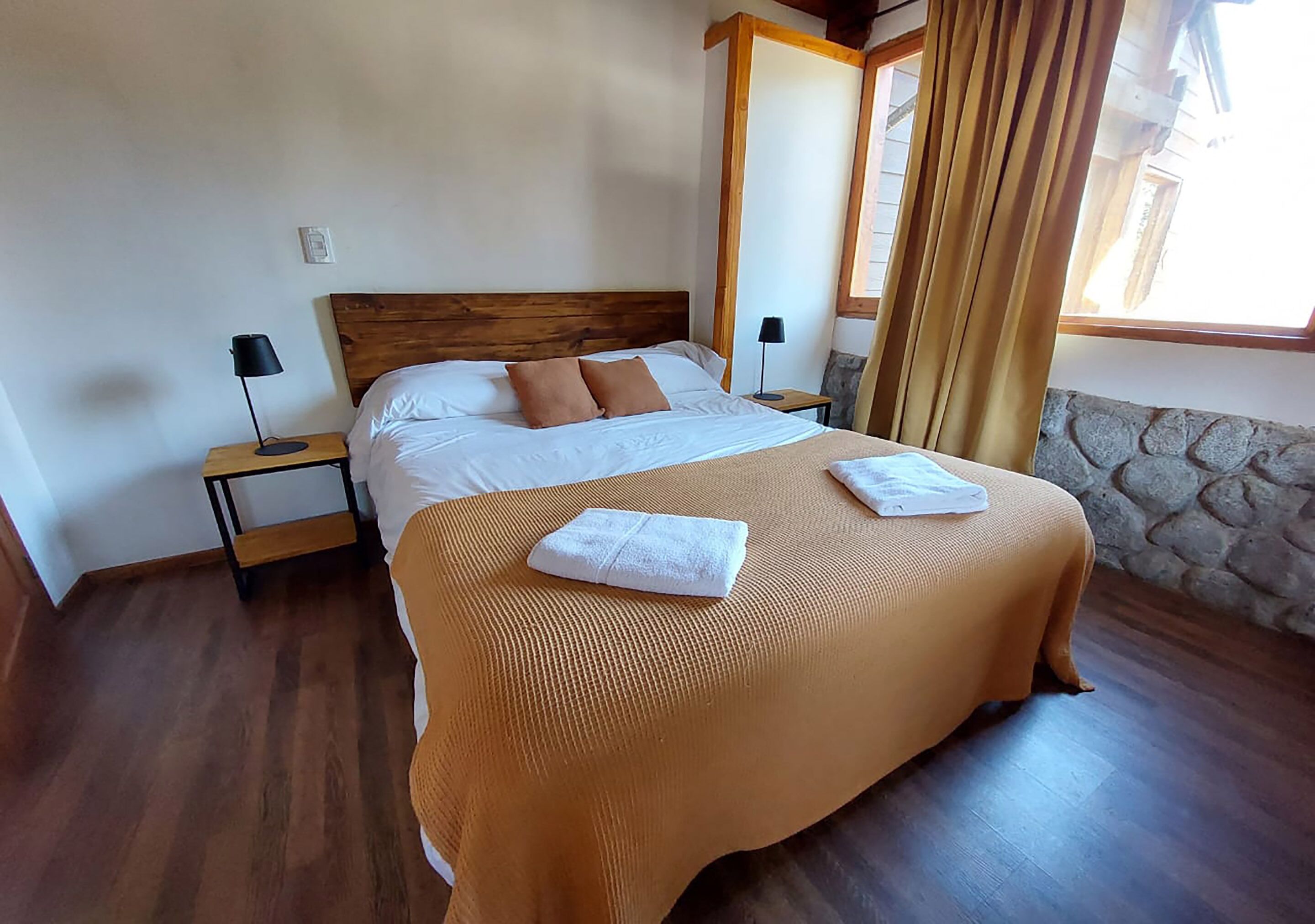 A neatly made bed with a wooden headboard, two bedside tables with lamps, a stone wall, and a window with curtains.
