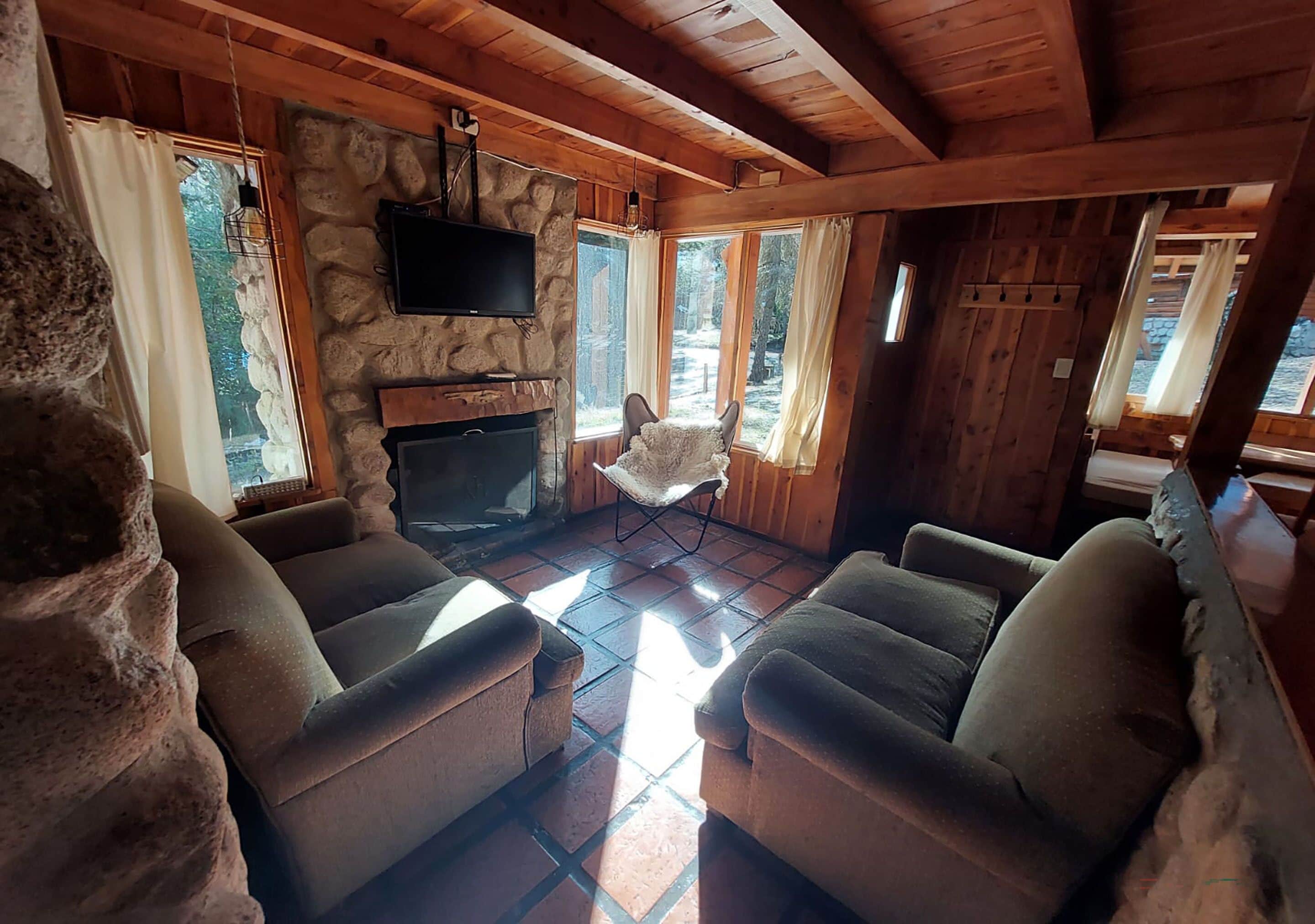 Interior acogedor de una cabaña con chimenea de piedra, paredes de madera y grandes ventanales con vistas al bosque.