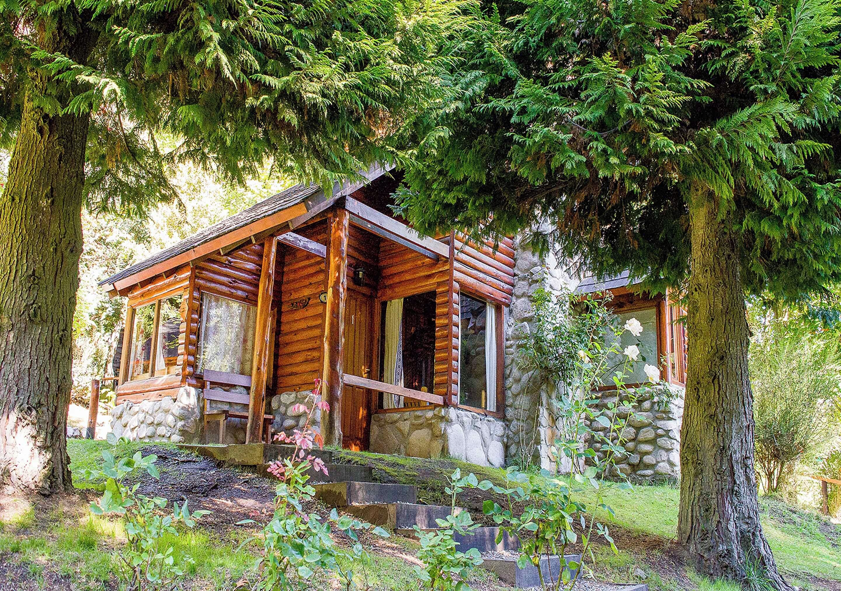 Une cabane rustique en rondins de bois, posée sur des fondations en pierre, entourée d’une végétation luxuriante et de grands arbres.