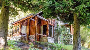 Une cabane rustique en rondins de bois, posée sur des fondations en pierre, entourée d’une végétation luxuriante et de grands arbres.