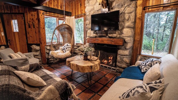 A cozy cabin interior with a stone fireplace, a television mounted on the wall, and a comfortable seating area with a woven chair and cushioned sofas. - Ruma Andina by DOT Cabana (Villa La Angostura)