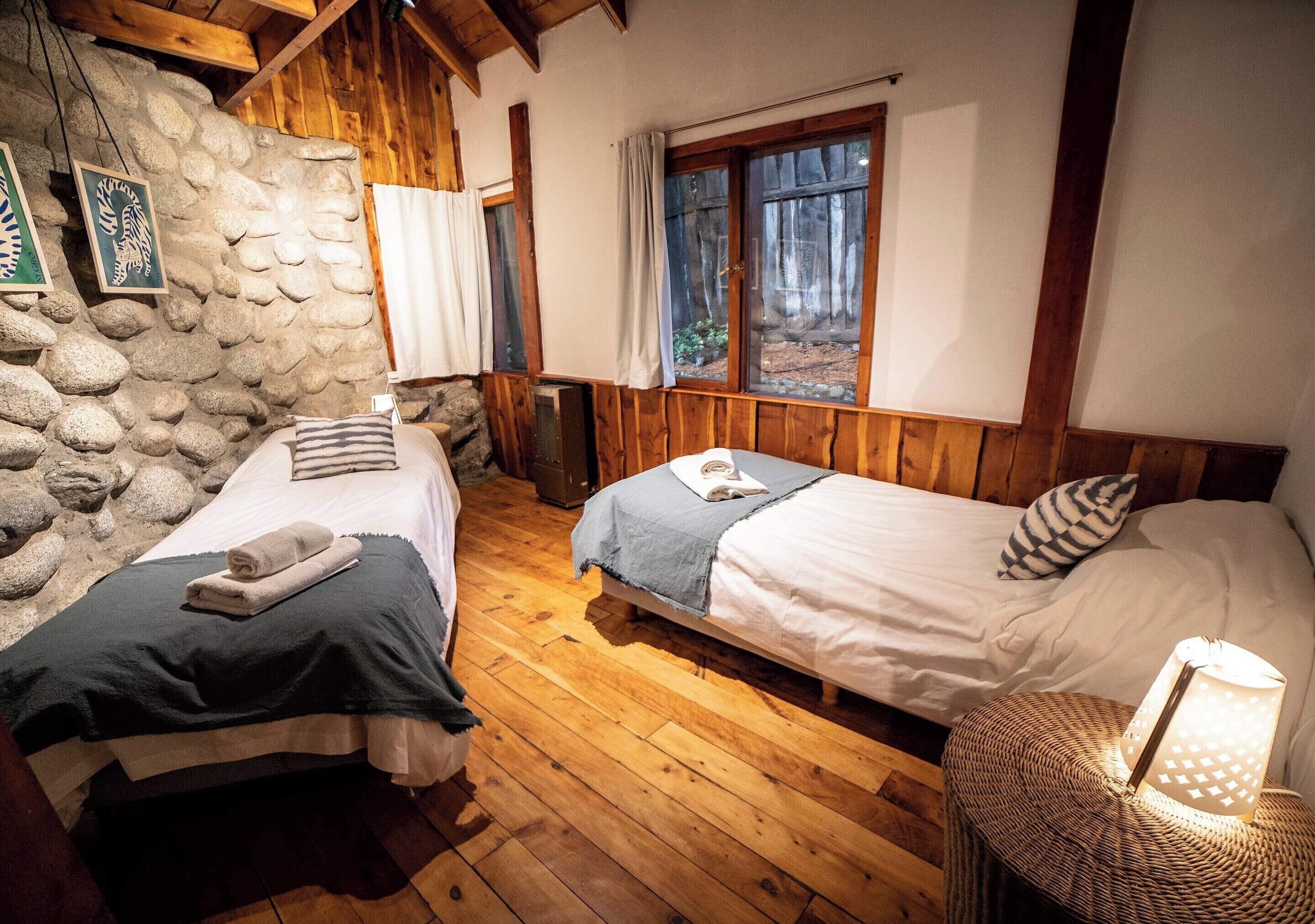 Una habitación rústica con dos camas, piso de madera, paredes de piedra y una ventana con cortinas.