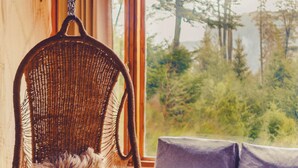 A wicker chair with a fluffy cushion, a grey sofa with two cushions, and a view of trees through a window. - Ruma Andina by DOT Cabana (Villa La Angostura)