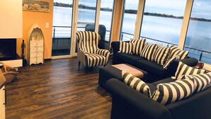 A living room with a black and white striped sofa, a striped armchair, and a wooden coffee table. A fireplace is visible on the left, and there are large windows offering a view of a lake. - Houseboat "ELANDI" - relaxation, fun & luxury (Bitterfeld)