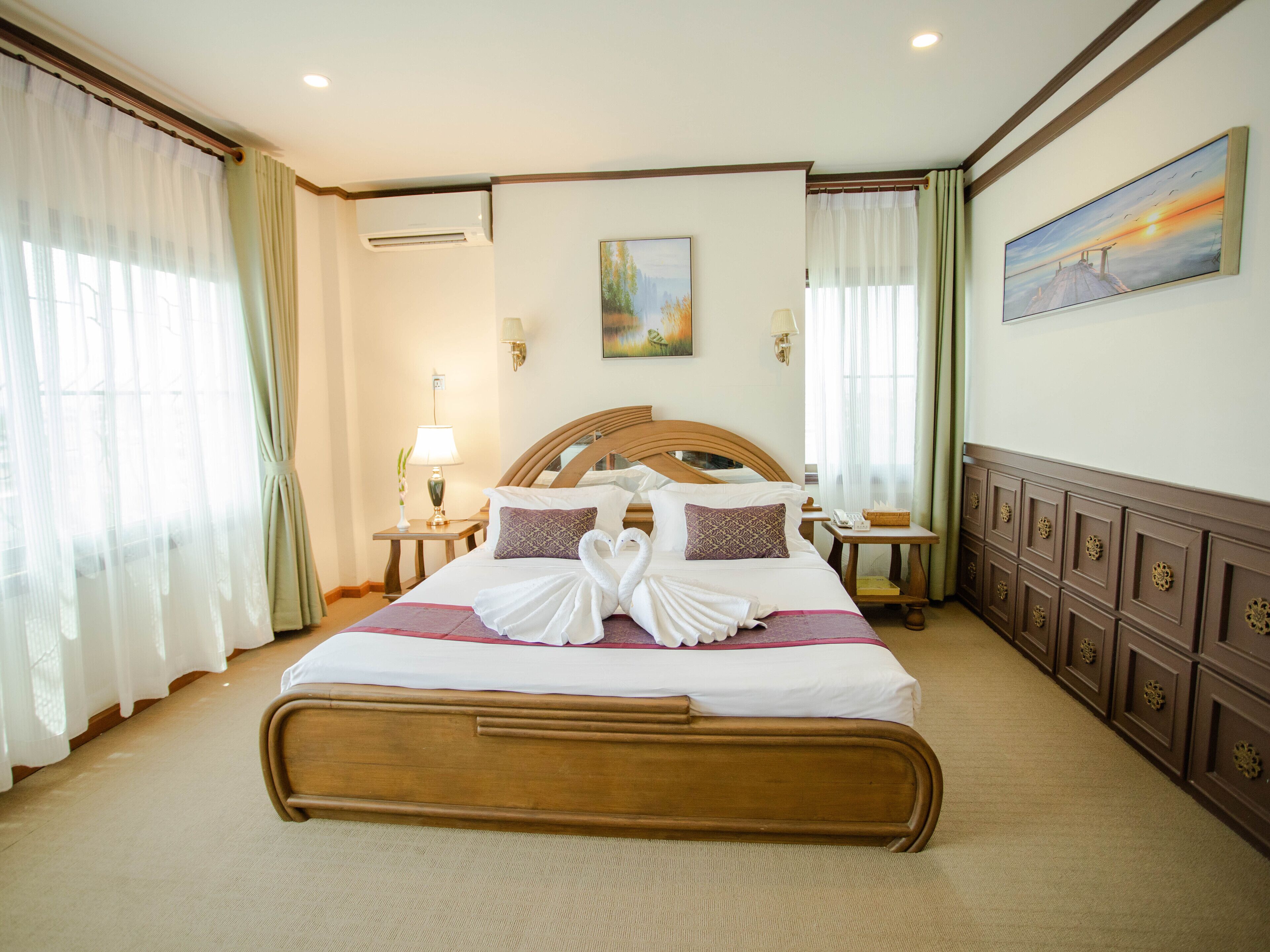 A hotel room with a large bed, two bedside tables, a wall-mounted air conditioner, and a painting above the bed.