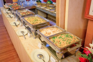 A buffet with various dishes in metal serving trays, arranged on a long table with plates and spoons. - E-Outfitting Golden Country Hotel (Mandalay)