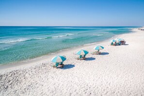 Una playa con arena blanca y agua azul cristalina. Varias sombrillas y sillas de playa azules y blancas a lo largo de la orilla.