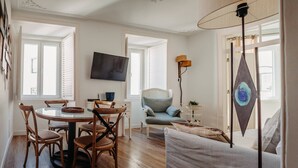 A living room with a round glass table, wooden chairs, a blue armchair, a white sofa, a floor lamp, and a TV mounted on the wall. - Bright and Spacious Apt on the City Centre (Lisboa)