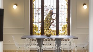 Un coin repas avec une table et quatre chaises en fil de fer blanc, un vitrail décoratif représentant un paon et des lampes fixées au mur.