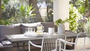 Un espace extérieur aménagé avec une table en osier, des chaises blanches, un banc rembourré et un pot de fleurs.