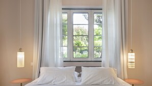 Un lit bien fait, avec du linge de lit blanc, une fenêtre avec des rideaux et deux lampes fixées au mur.