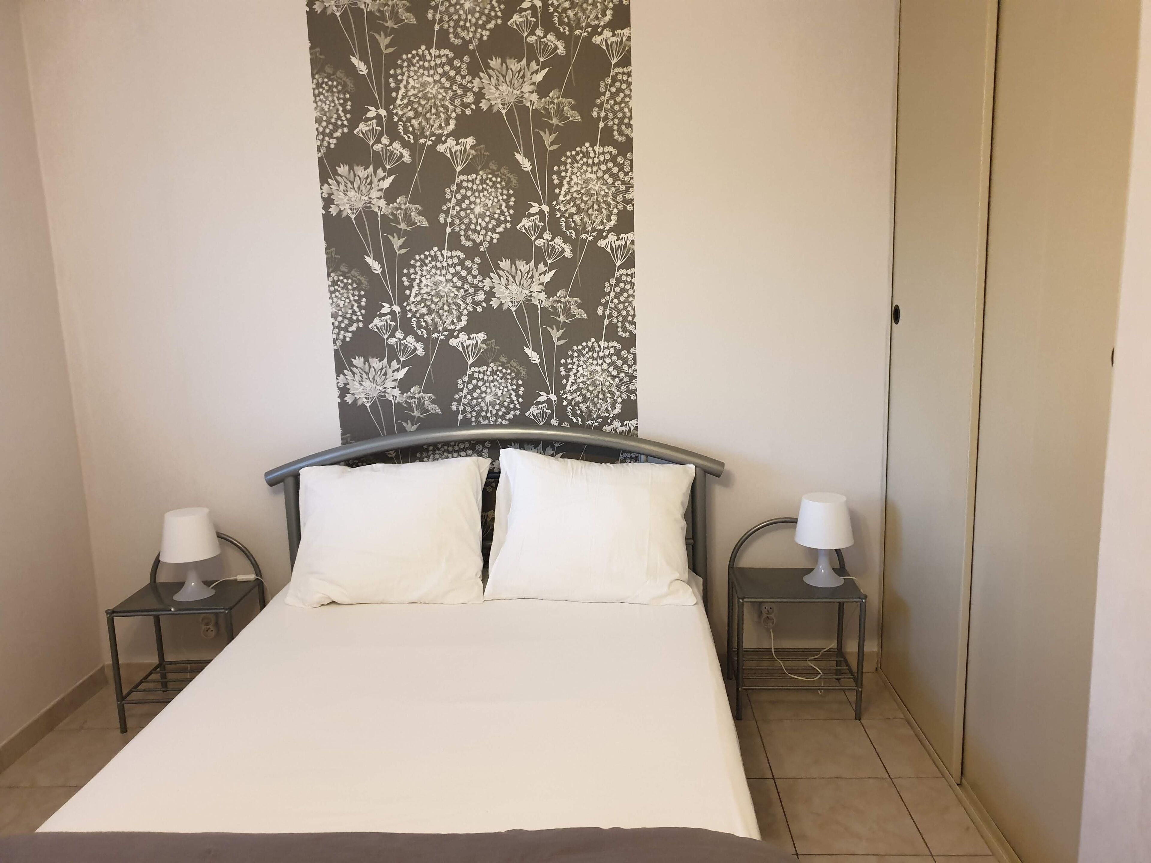 Een slaapkamer met een bed, twee nachtkastjes, een lamp op elk kastje, en een behang met bloemenpatroon boven het bed.