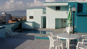 A rooftop pool area with white chairs and tables, a green umbrella, and a modern building with a blue roof in the background. - Crystal Apart Hotel (Macaé)