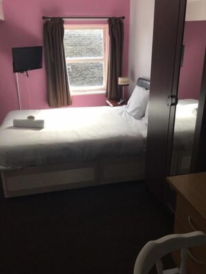 A single bed with a headboard, a bedside table, a lamp, and a window with curtains. - Gateway Lodge (Cleckheaton)