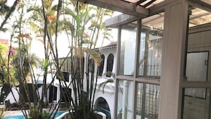 A covered outdoor area with a pool, potted plants, and a white building with arched windows. - Skina Comfort - Stays & Events - Large House with Pool! (Guarujá)