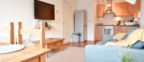A modern apartment with a dining area, a living room with a sofa, a kitchen with wooden cabinets, a TV mounted on the wall, and a small table with a plant.