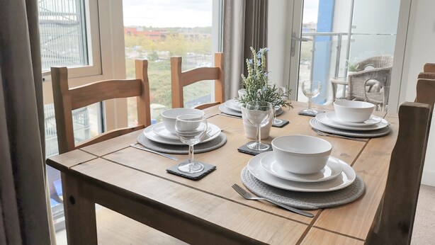 A dining table set with white plates, bowls, and wine glasses, surrounded by wooden chairs. - MK Hub Apartment with Oak Furniture (Milton Keynes)