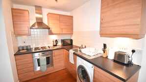 A compact kitchen with wooden cabinets, a washing machine, a toaster, and a microwave. - MK Hub Apartment with Oak Furniture (Milton Keynes)