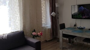 Una sala de estar con un sofá, una mesa de comedor con sillas, un televisor montado en la pared y un ventanal con cortinas.