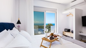 A modern hotel room with a large bed, a bedside table with a tray of breakfast, a TV, and a balcony with a view of the sea. - Evilio Suites, Inspired Family Retreat with Pool (Prinos, Rethymno)