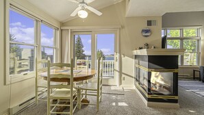 A room with a fireplace, a dining table with chairs, a ceiling fan, and large windows with a view of the outdoors. - Second Floor Lakeview Condo (Manistee)