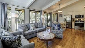 Een ruime woonkamer met een grijze bank, gestreept fauteuils en een ronde salontafel. Achtergrond zichtbaar van een keuken met roestvrijstalen apparatuur.