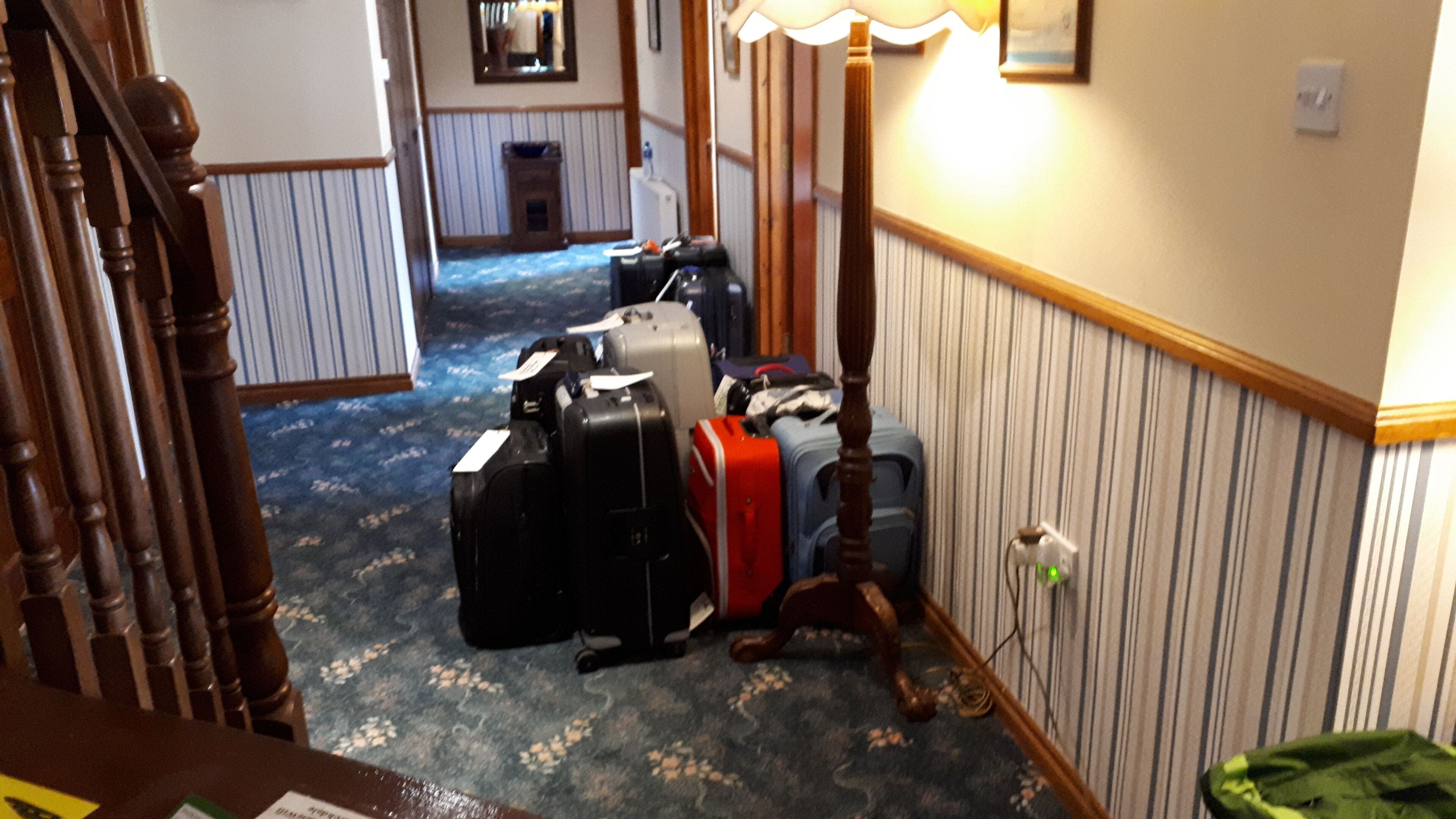 A hallway with luggage stacked against the wall, a wooden staircase, and a carpeted floor.