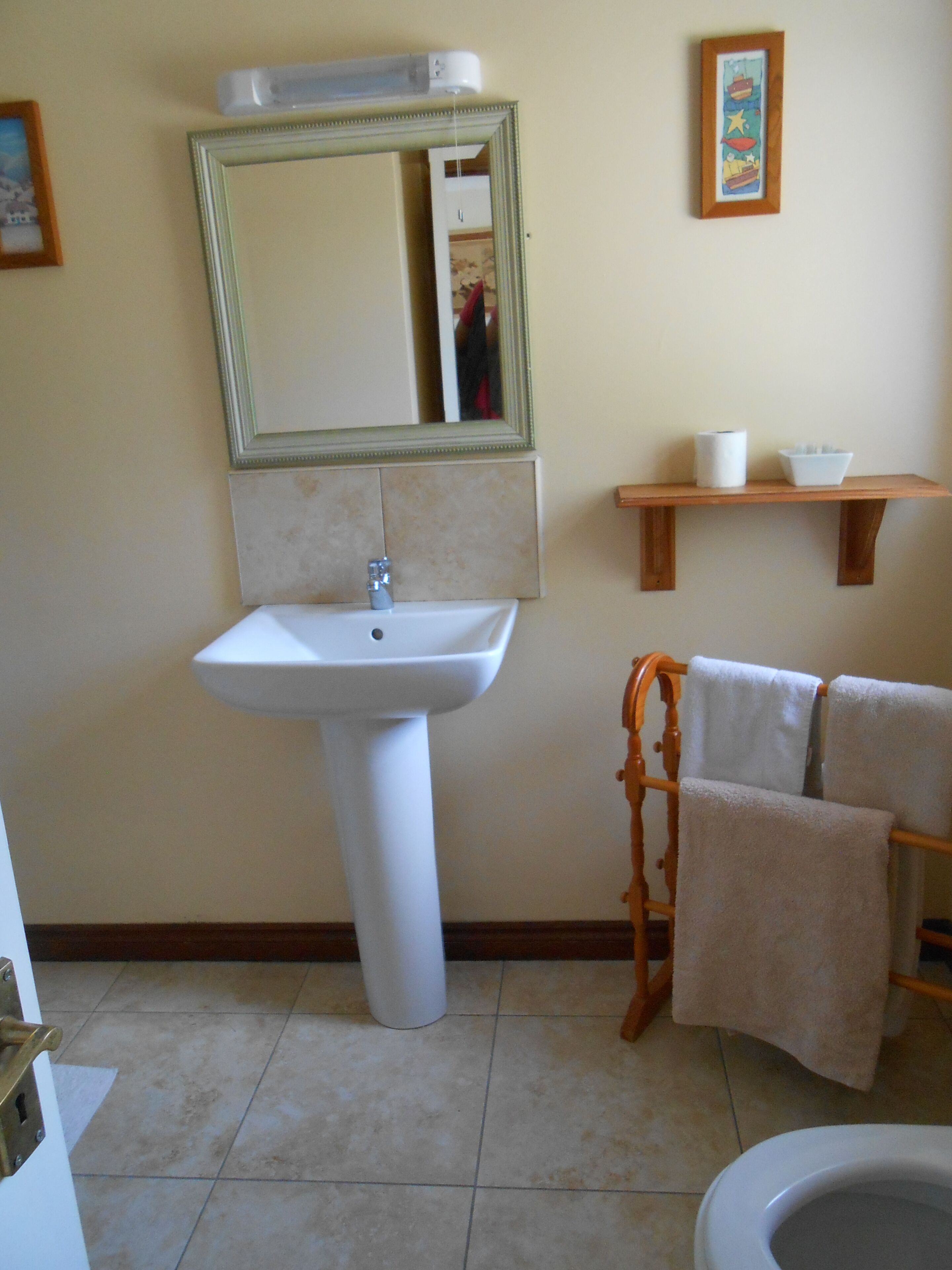 A bathroom with a pedestal sink, a wooden towel rack, a toilet, and a shelf with a bowl and a roll of paper towels.
