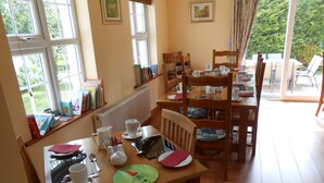 A dining area with a wooden table set for four, surrounded by chairs. There are books on a shelf to the left, and a window with a view of greenery outside. - Birchdale House B&B (Greenan)