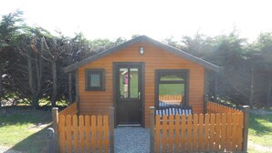 A small wooden cabin with a door and windows, surrounded by a wooden fence, set in a grassy area with trees in the background. - Birchdale House B&B (Greenan)