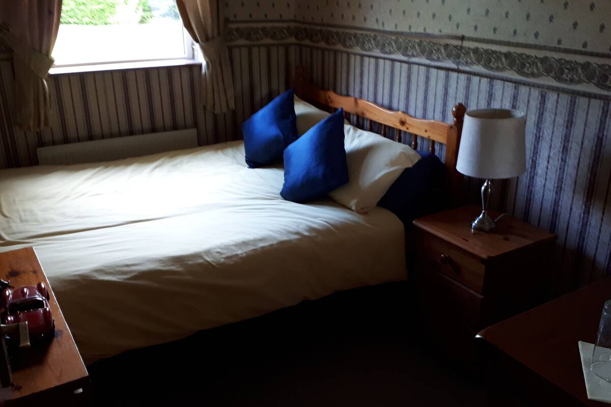 Ein Bett mit weißer Bettwäsche und blauen Kissen, ein Nachttisch mit Lampe sowie ein kleiner Tisch mit Wasserflasche und Gläsern.