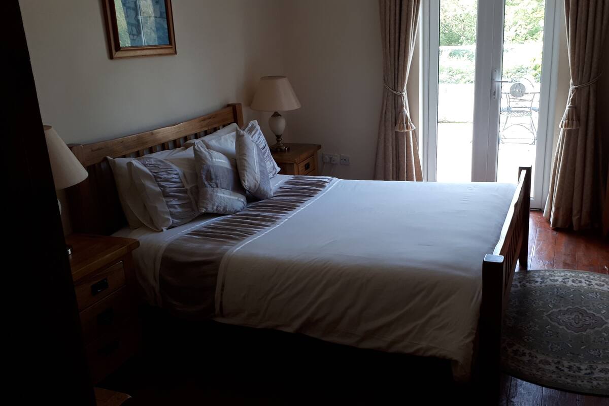 Ein ordentlich bezogenes Bett mit Holzkopfstütze, ein Nachttisch mit Lampe, eine Kommode mit Schubladen und ein Blick ins Freie durch ein Fenster.