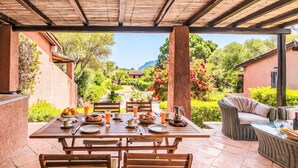 Una terrazza esterna con tavolo di legno apparecchiato per la colazione, circondata da mobili in vimini e vista su un giardino e delle colline.