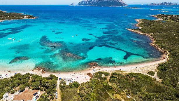 Una vista aerea di una zona costiera con spiaggia, imbarcazioni e un'isola rocciosa in lontananza.
