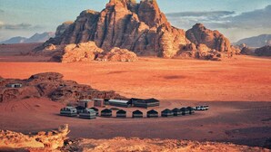 A desert landscape with a large rock formation in the background and a row of tents in the foreground. - Ultimate (Wadi Rum)