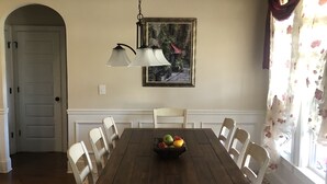 En spisestue med et træbord, hvide stole, en lysekrone, et indrammet billede og blomstret gardin.