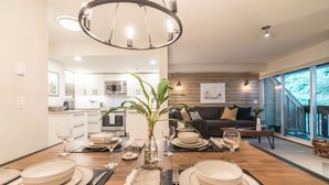 A modern dining area with a wooden table set for a meal, a kitchen in the background, and a large window offering a view of the outdoors.