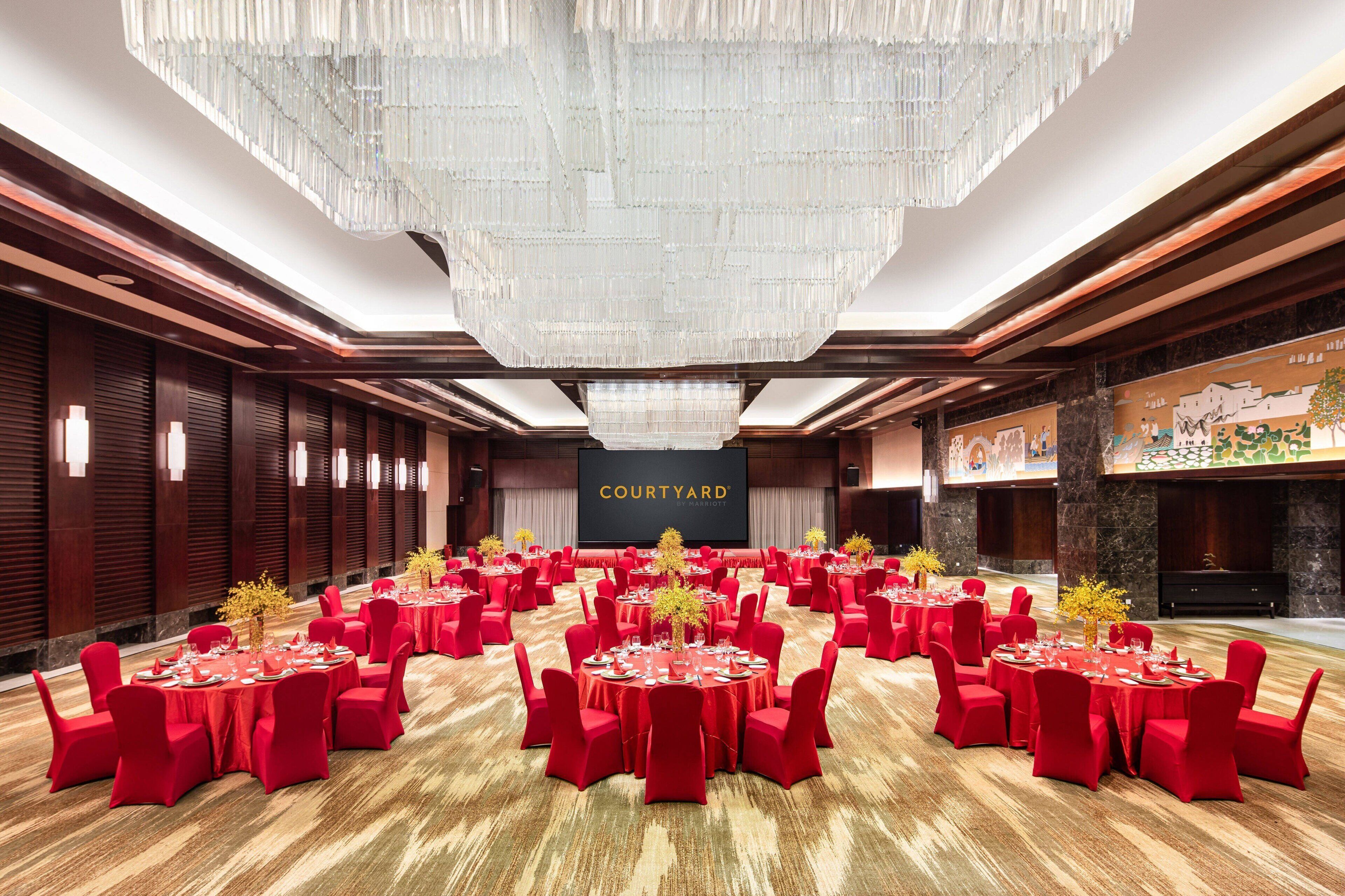 A large, well-lit conference room with multiple round tables, red chairs, and a chandelier.