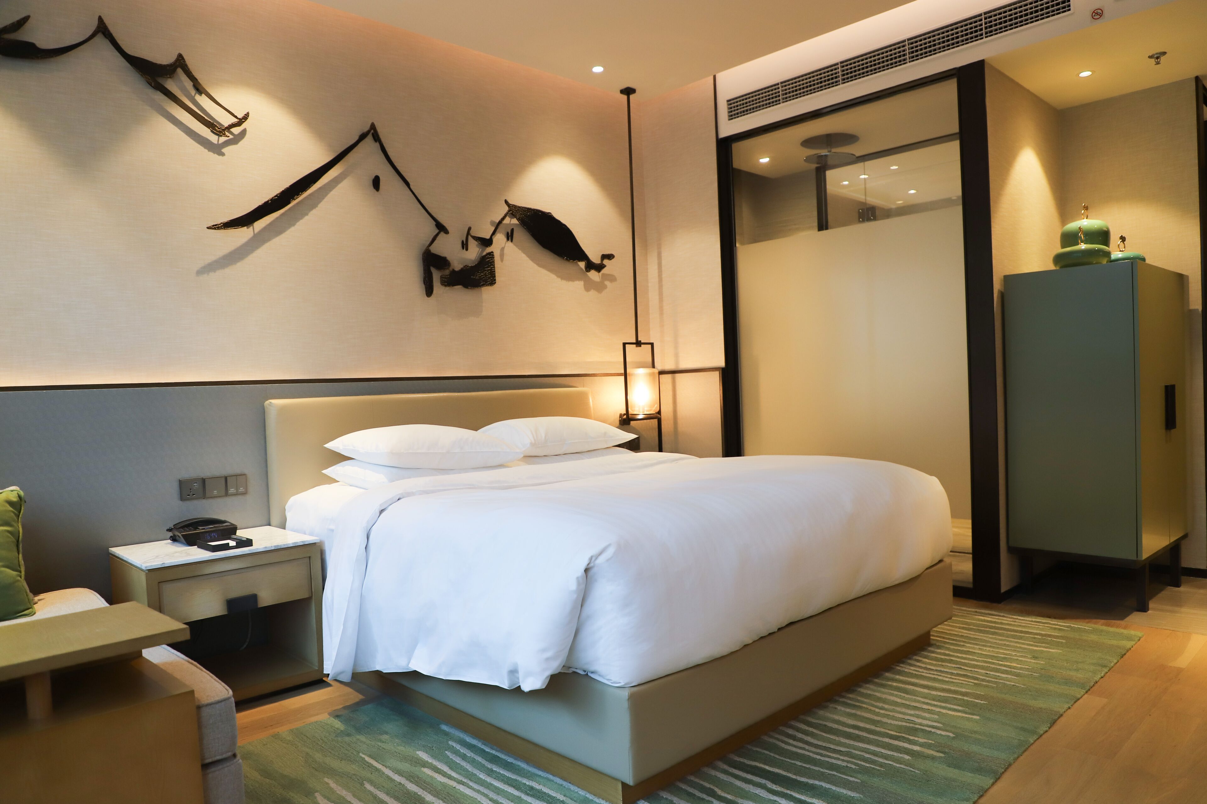 A modern hotel room with a large bed, a bedside table, a nightstand, a green rug, and a wall-mounted artwork.