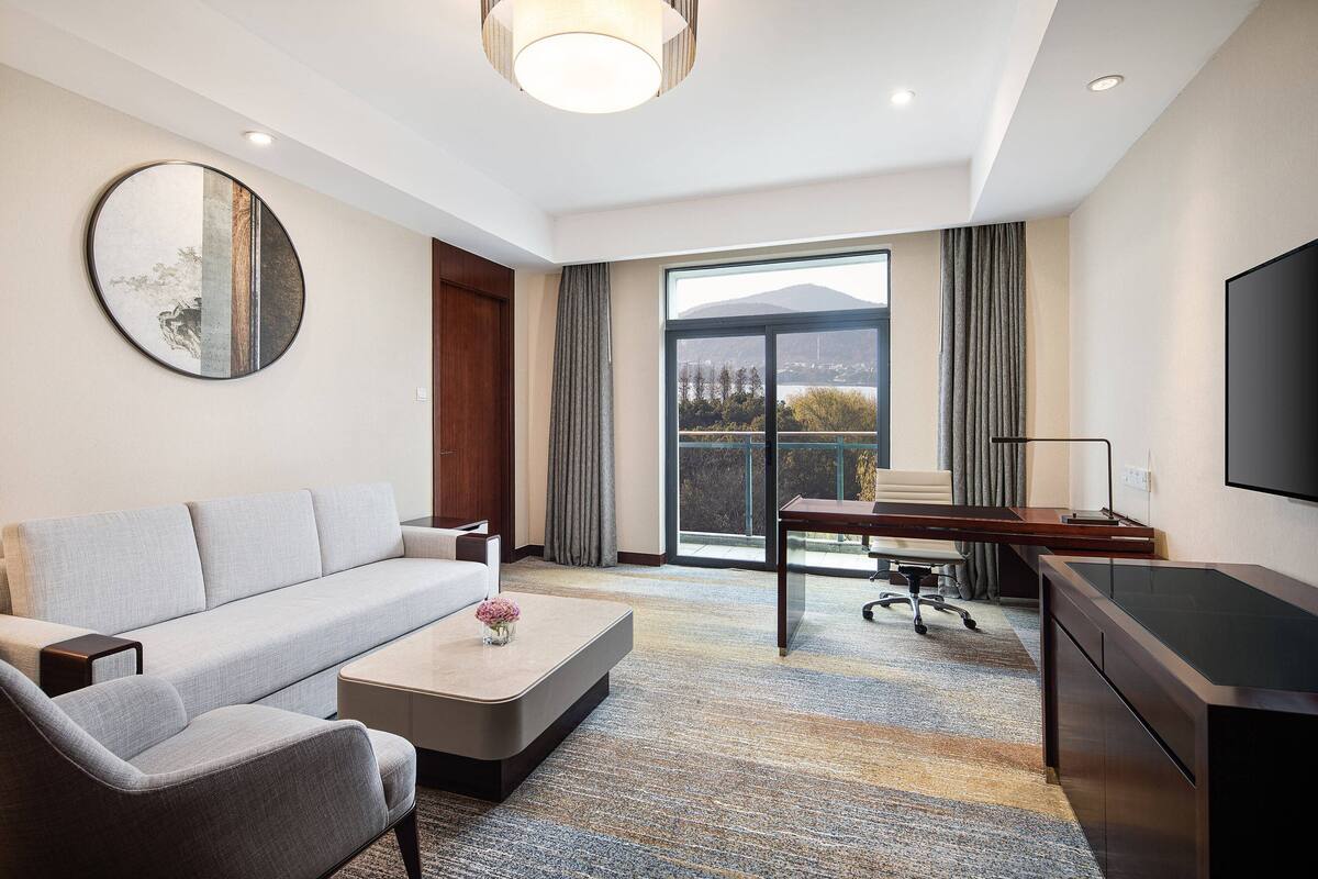 Una habitación de hotel moderna con sofá, escritorio, televisión de pantalla plana y un amplio ventanal con vista a la ciudad.