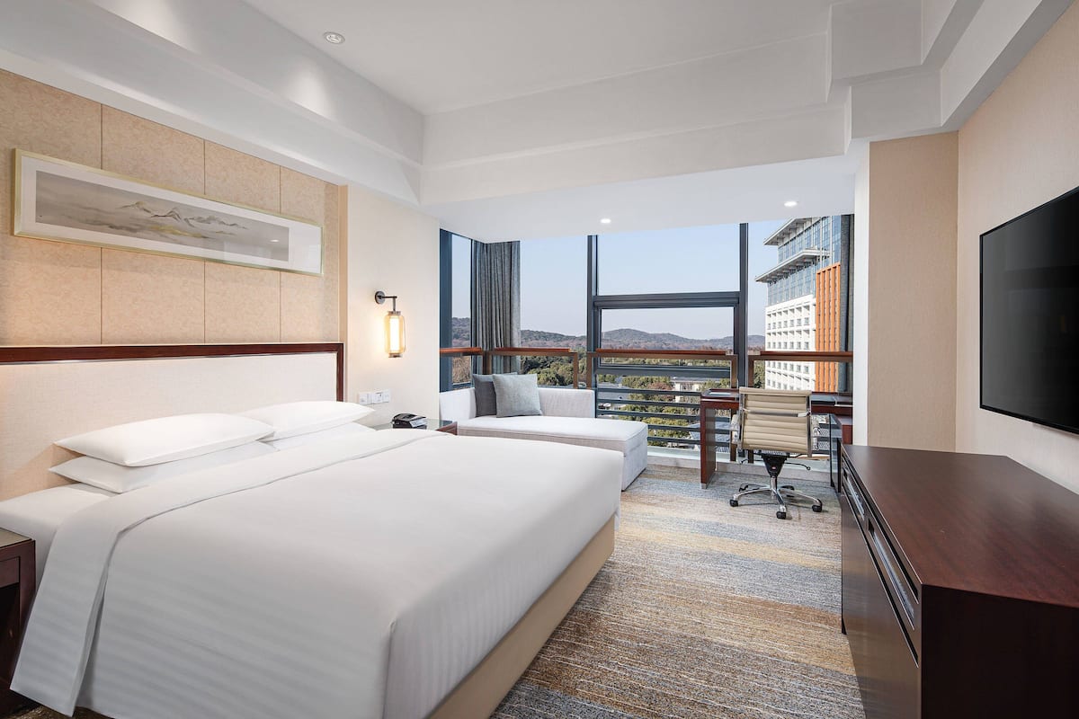 Habitación de hotel con una cama grande, un escritorio, una silla y un televisor. Se ve una ventana con vistas a edificios y colinas.