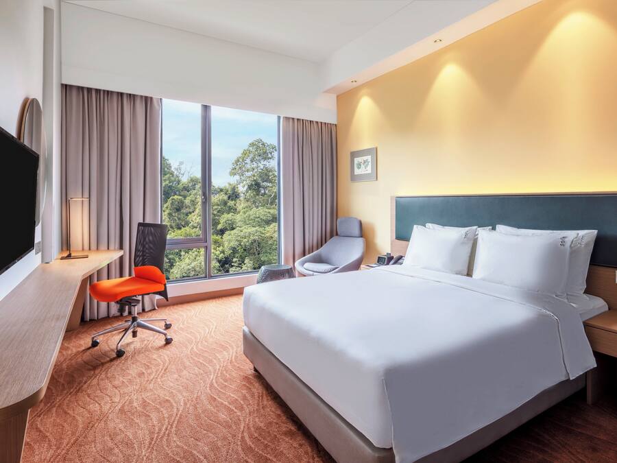 Ein Hotelzimmer mit einem großen Bett, einem Schreibtisch mit Fernseher, einem Sessel und Blick auf Grünanlagen durch das Fenster.