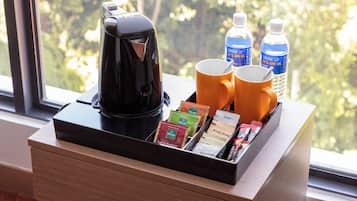 A coffee maker, two mugs, water bottles, and a tray with tea and snack packets on a wooden table by a window.
