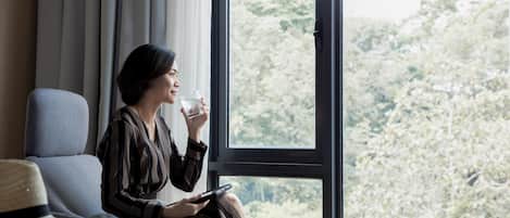 A person sitting by a window with a view of trees, holding a glass and a tablet.